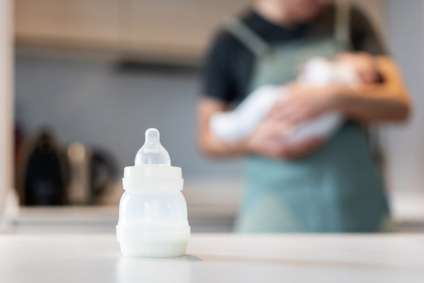 Close-up view of unrecognizable single dad with baby at kitchen counter feeding milk bottle to newborn.