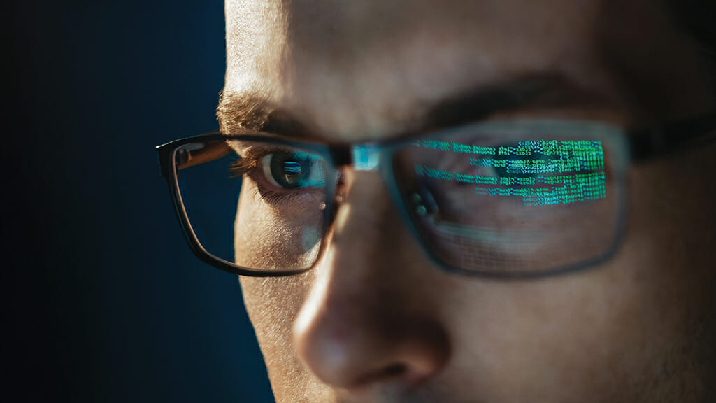 Close-up portrait of focused software engineer wearing eyeglasses looking at computer screen working with big data. Programming code reflecting in glasses. Data science, Machine Learning, AI concept
